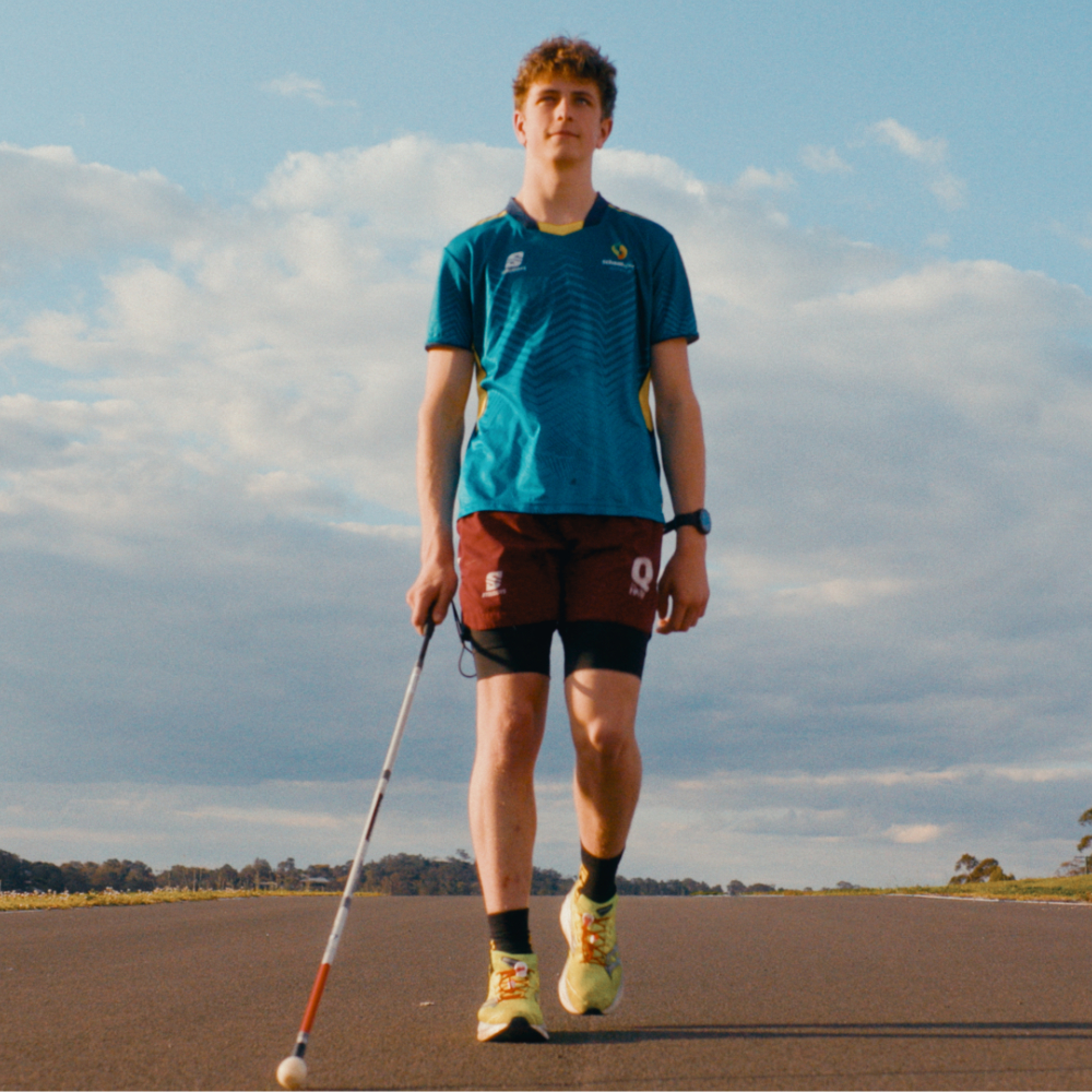 Mitchell walks down a road, holding a white cane in his right hand. He wears a teal t-shirt, maroon shorts, and bright yellow sneakers against a cloudy sky.