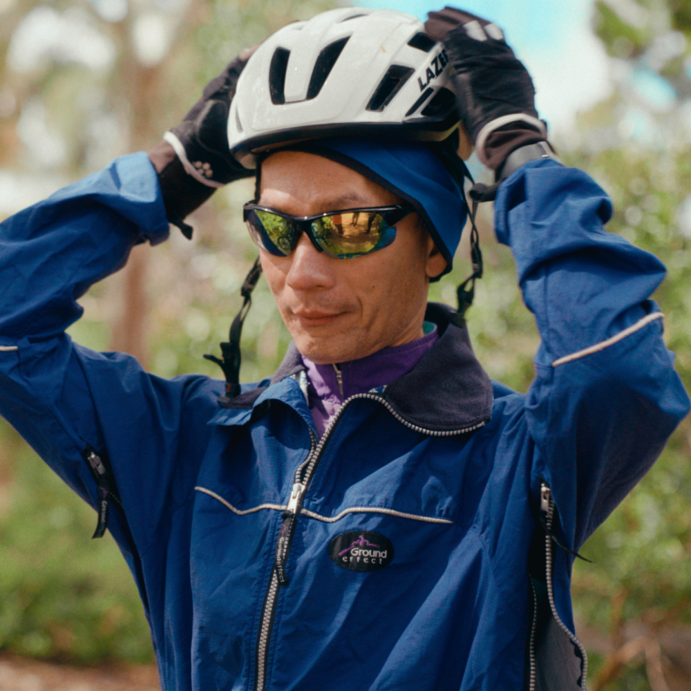 Simon adjusts his white bicycle helmet outdoors, wearing a blue jacket, sunglasses, and cycling gloves.