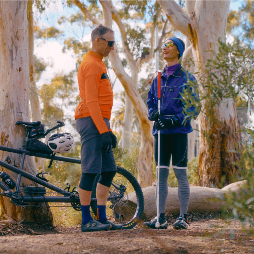 Two men stand smiling among eucalyptus trees next to a tandem mountain bike, with Simon on the right holding a white cane.