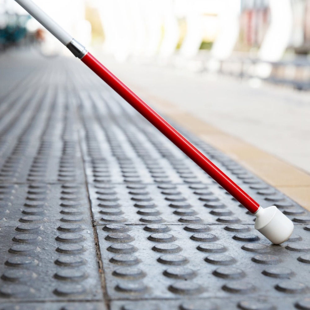A red-and-white mobility cane sweeps across tactile paving tiles on a train platform.