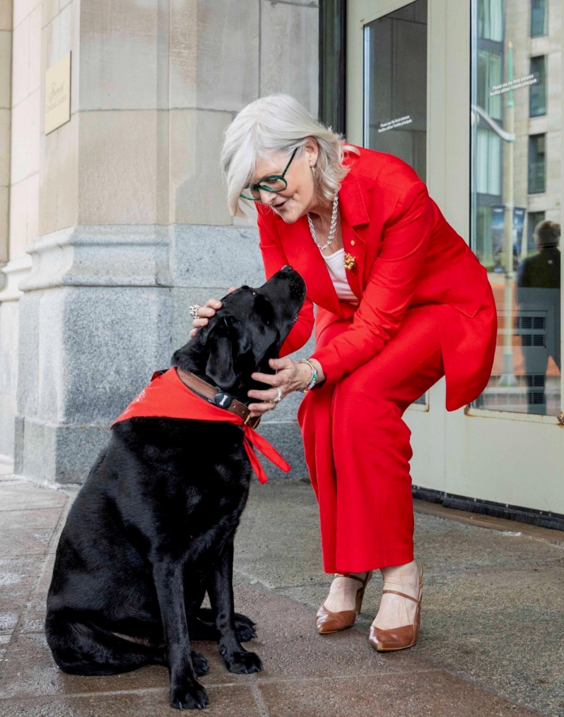 A woman with short grey hair and glasses, wearing a bright red pantsuit, kneels down and affectionately pets a black Labrador wearing a red bandana.